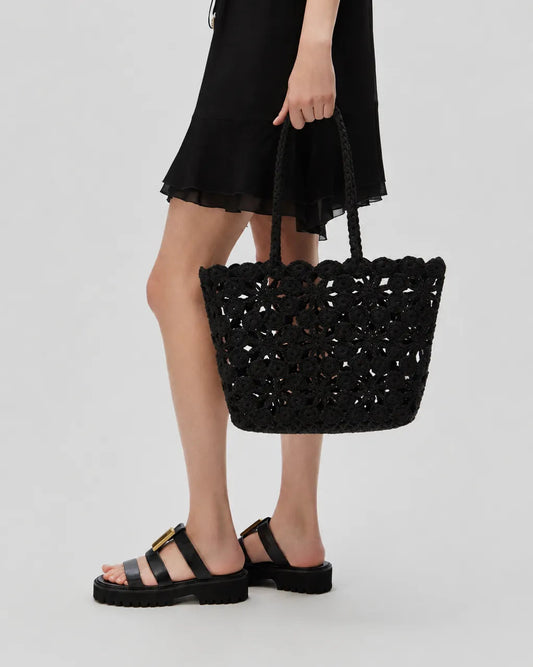 A woman wearing a black dress and black slide sandals holds the black crocheted tote basket bag against a light gray studio background.