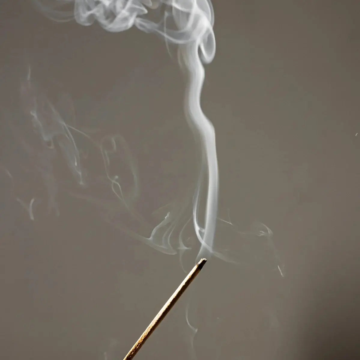 A single incense stick burning, with a curl of white smoke rising against a grey background.