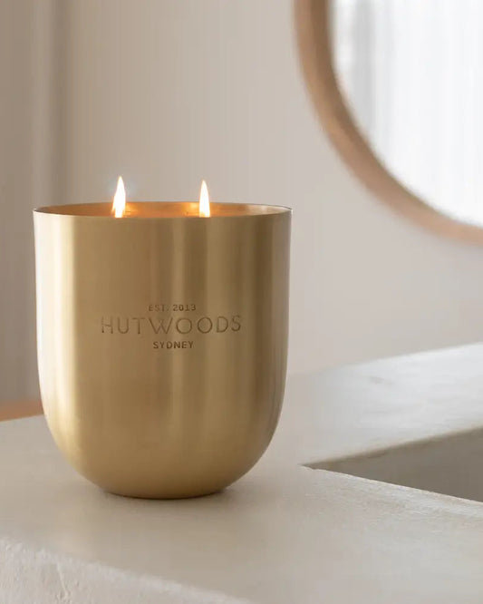 A lit gold Hutwoods Extra Large gold candle burning with two wicks on a benchtop in a calm room