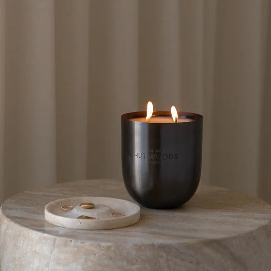 A lit Hutwoods Bergamot & Anjou Pear Candle in brushed black vessel on a marble side table