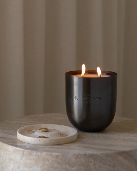 A lit Hutwoods Bergamot & Anjou Pear Candle in brushed black vessel on a marble side table