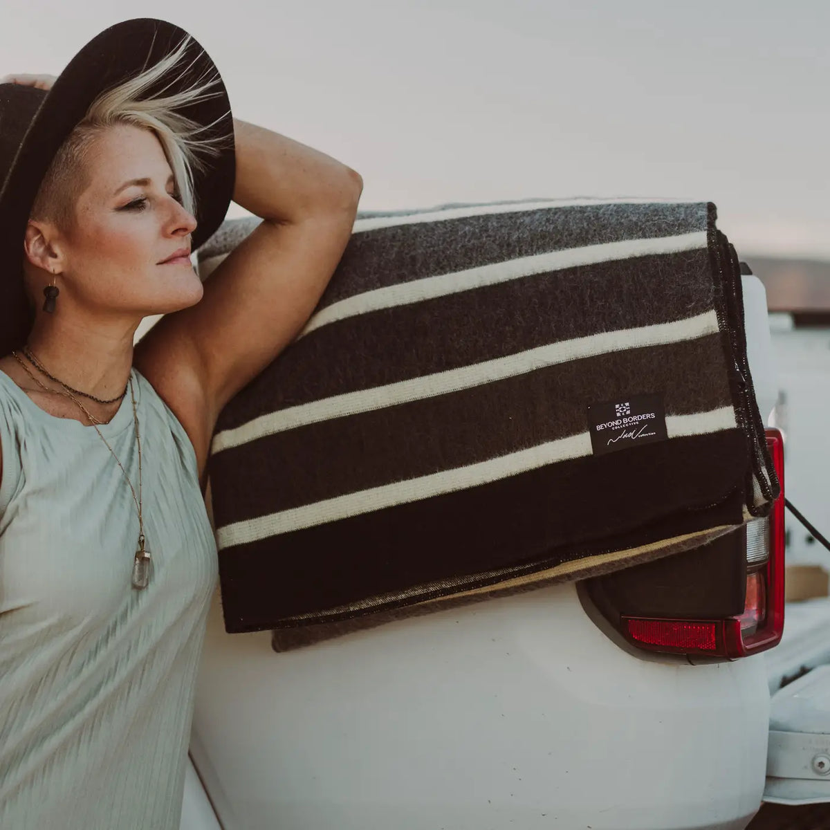 Woman in a wide-brim hat leaning on a ute with the striped Caminante blanket folded over the tailgate
