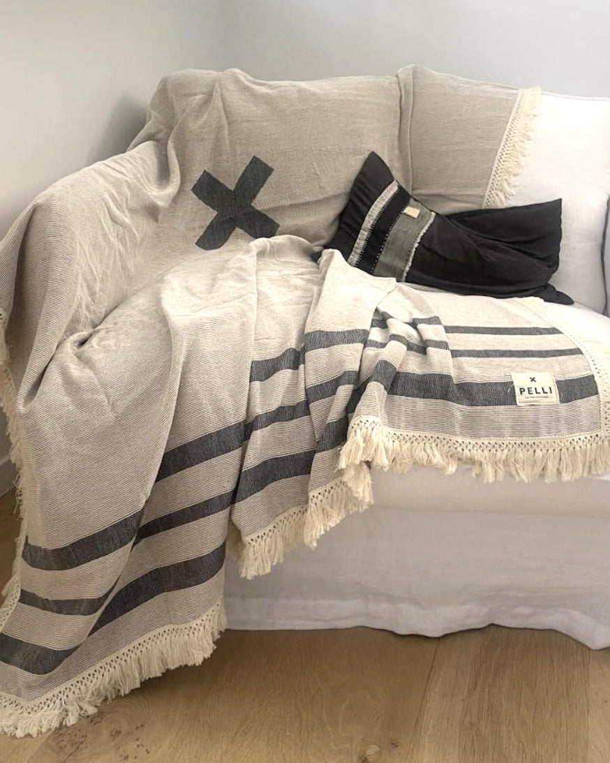 X Marks the Spot picnic blanket in charcoal by Pelli laid across a white couch with a black cushion on top. Looks comfy.