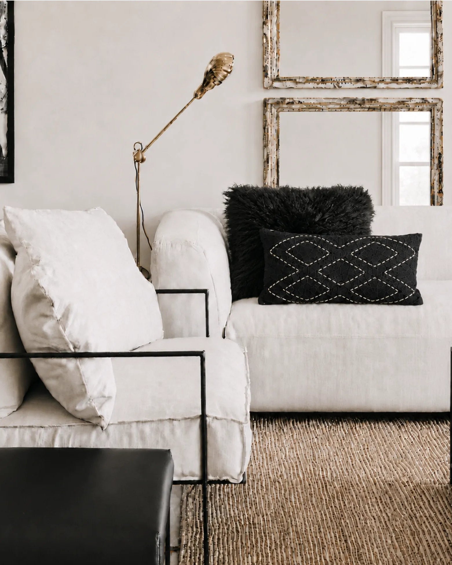 A styled living room corner featuring a white sofa with white and black throw pillows, a black metal side table, a gold floor lamp, and a woven jute rug against a neutral wall with two mirrors.