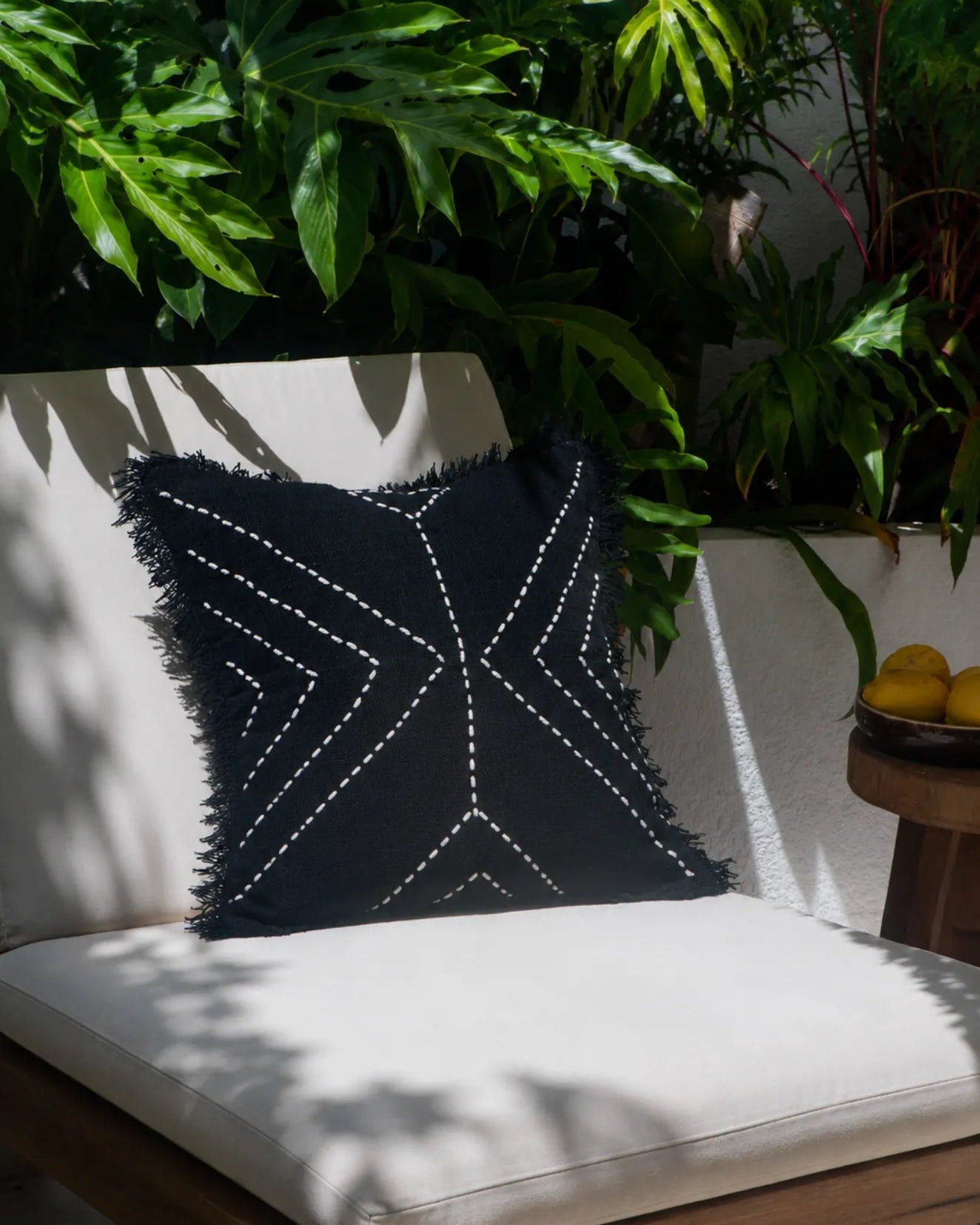 A square black throw pillow with white embroidered geometric patterns and fringed edges, resting on a white outdoor lounge chair surrounded by lush green plants.