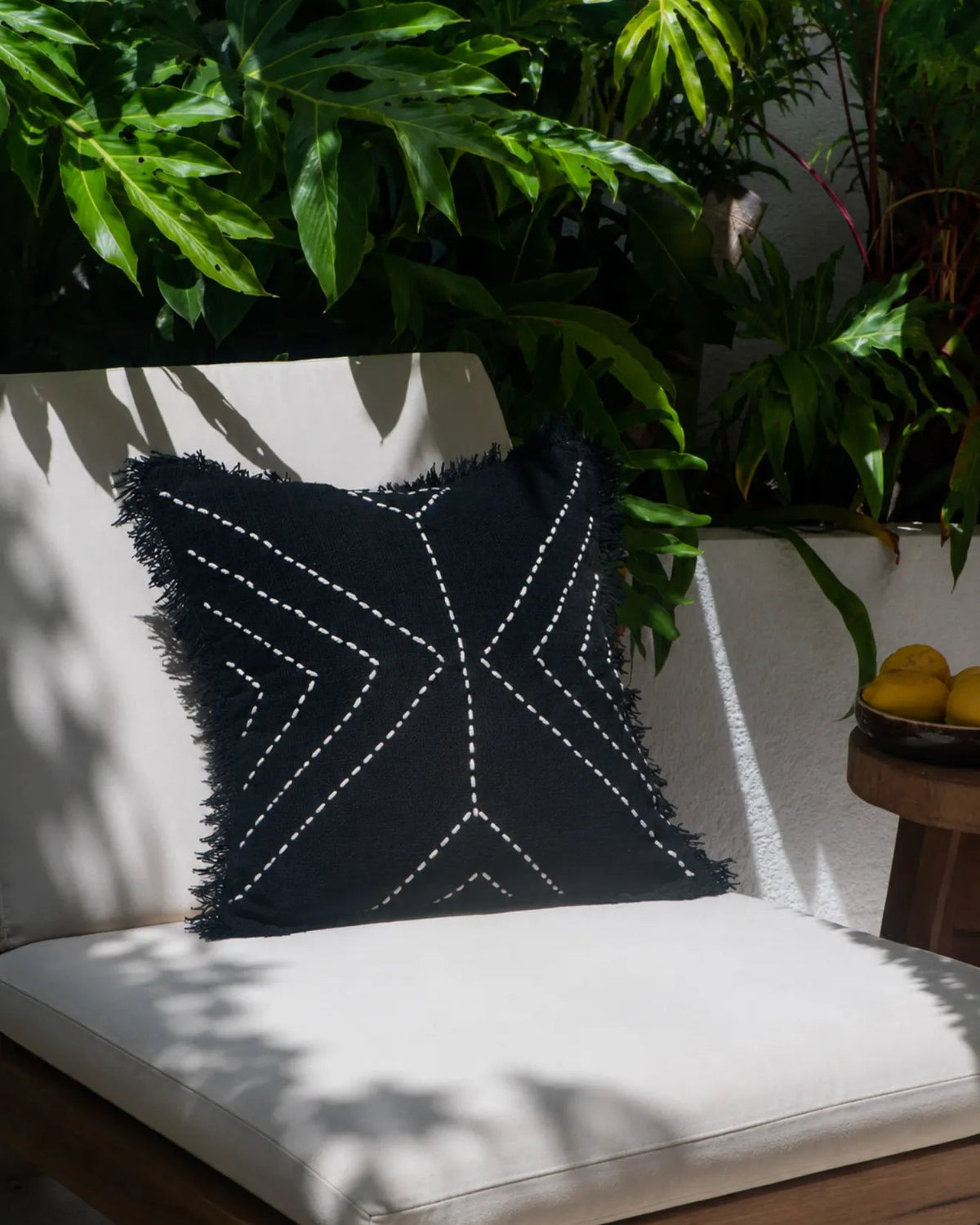 A square black throw pillow with white embroidered geometric patterns and fringed edges, resting on a white outdoor lounge chair surrounded by lush green plants.