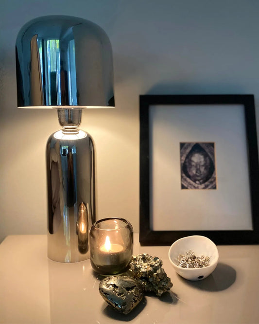 A polished silver metal table lamp with a rounded dome shade sits on a bedside table next to a lit candle in a glass holder, a small white bowl, and two pyrite stones the heart and raw, with a framed print leaning behind it against the wall.