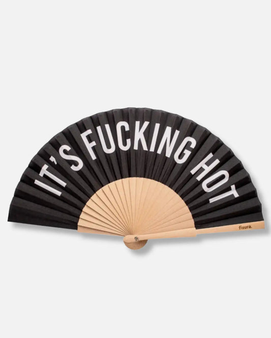 Black fabric hand fan with white lettering reading It's Fucking Hot, pale wooden ribs and a black cord loop, on a pale grey background.