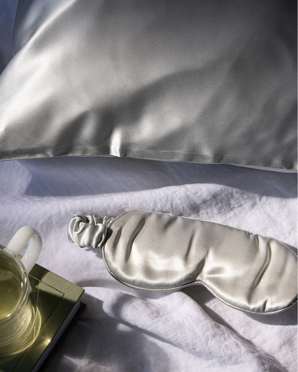 Gifts for the Hard-to-Buy-For: A luxurious grey silk eye mask lying on a white surface next to a cup of tea, with a pillow in the background. A great gift for anyone who values sleep.