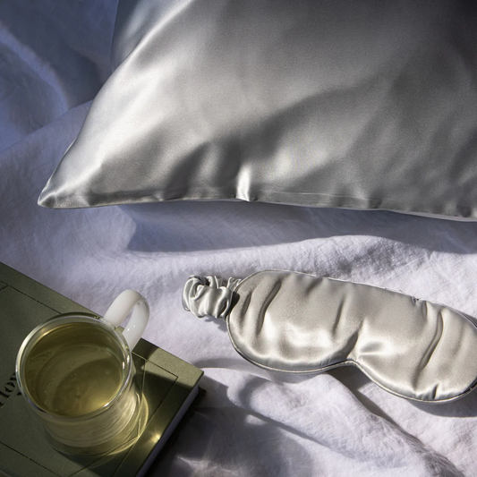 A luxurious grey silk eye mask lying on a white surface next to a cup of tea, with a pillow in the background.