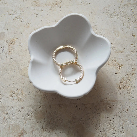 A white flower-shaped trinket bowl made from recycled resin, containing a pair of gold-toned hoop earrings, placed on a textured surface.