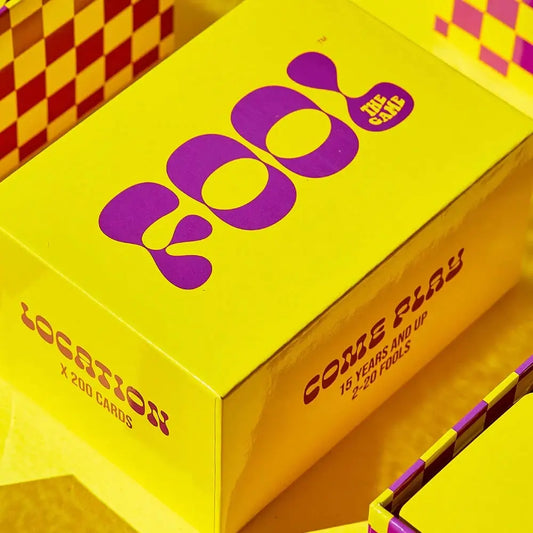 The image shows a brightly colored box of the card game 'Fool the Game' with a yellow background and purple text, featuring the game's title and a brief description.