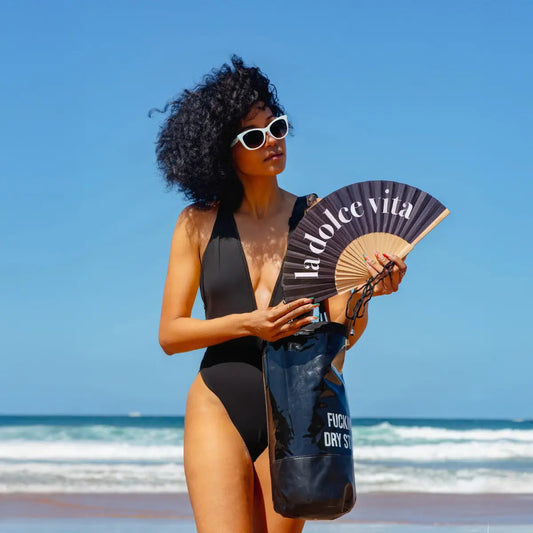 Woman on a beach holding a 'la dolce vita' fan with ocean and clear sky in the background