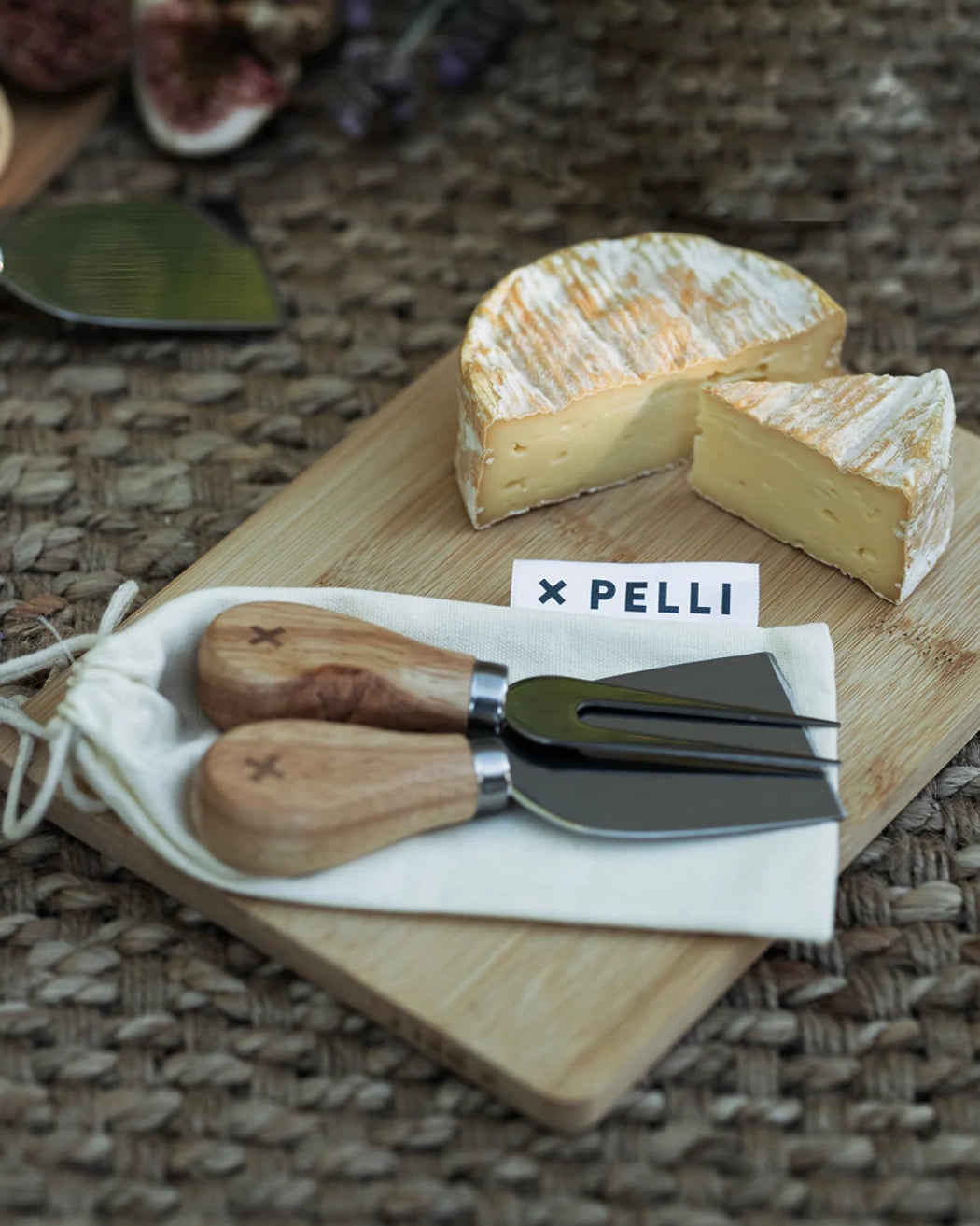 The Pelli Cheeseboard set with a wedge of cheese, two cheese knives, and a Pelli branded pouch on a woven mat.
