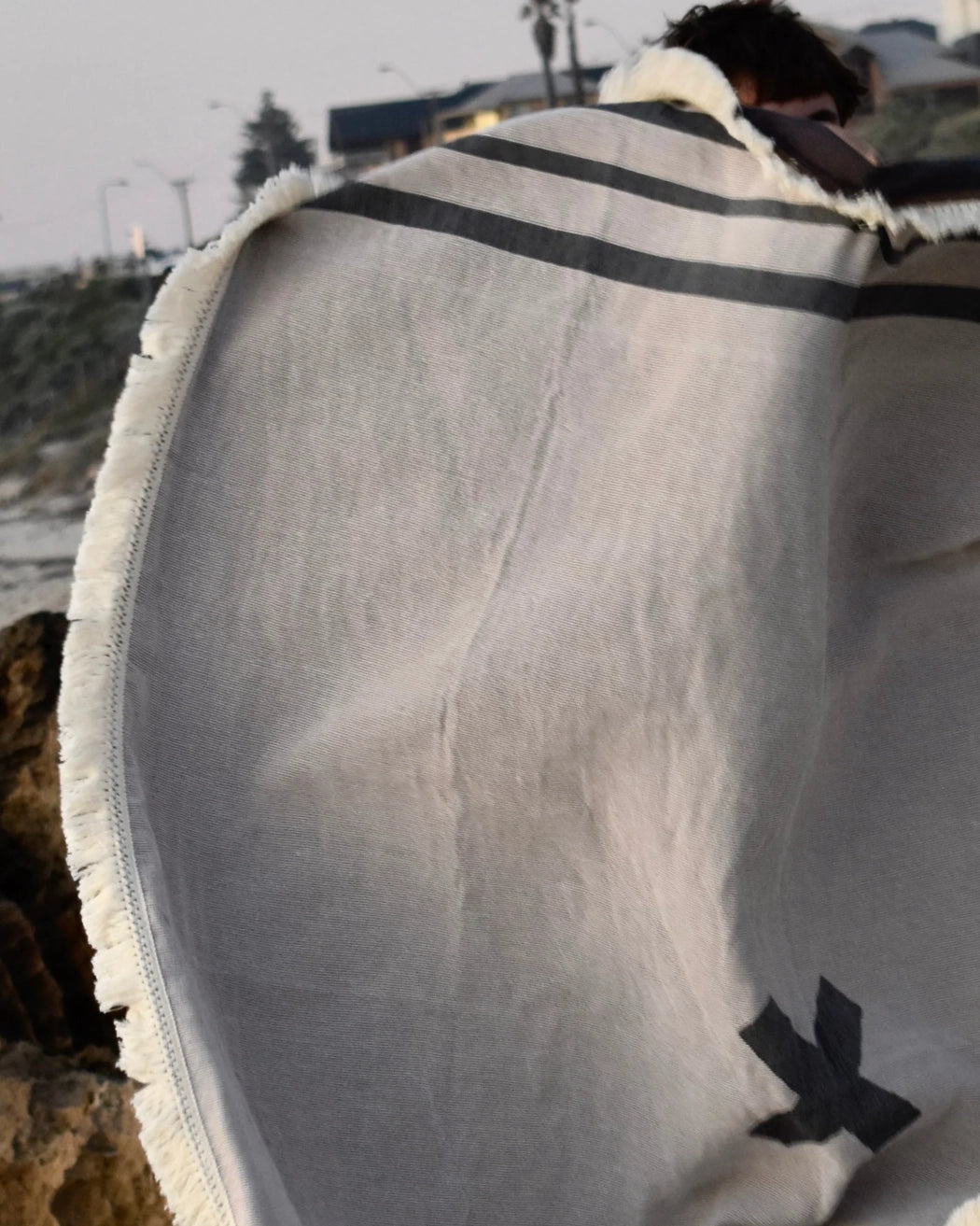 X Marks the Spot picnic blanket in charcoal by Pelli held up by someone standing on the beach as they fluff up the blankie to put it on the ground