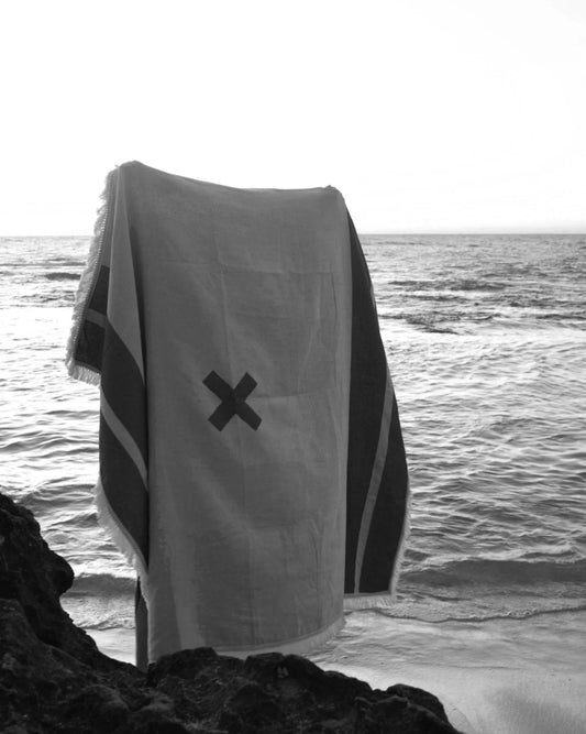 X Marks the Spot picnic blanket in charcoal by Pelli held up by someone standing on the beach