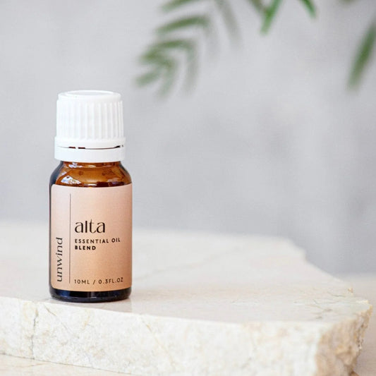 Bottle of Alta UNWIND essential oil blend on a white surface with a blurred background.