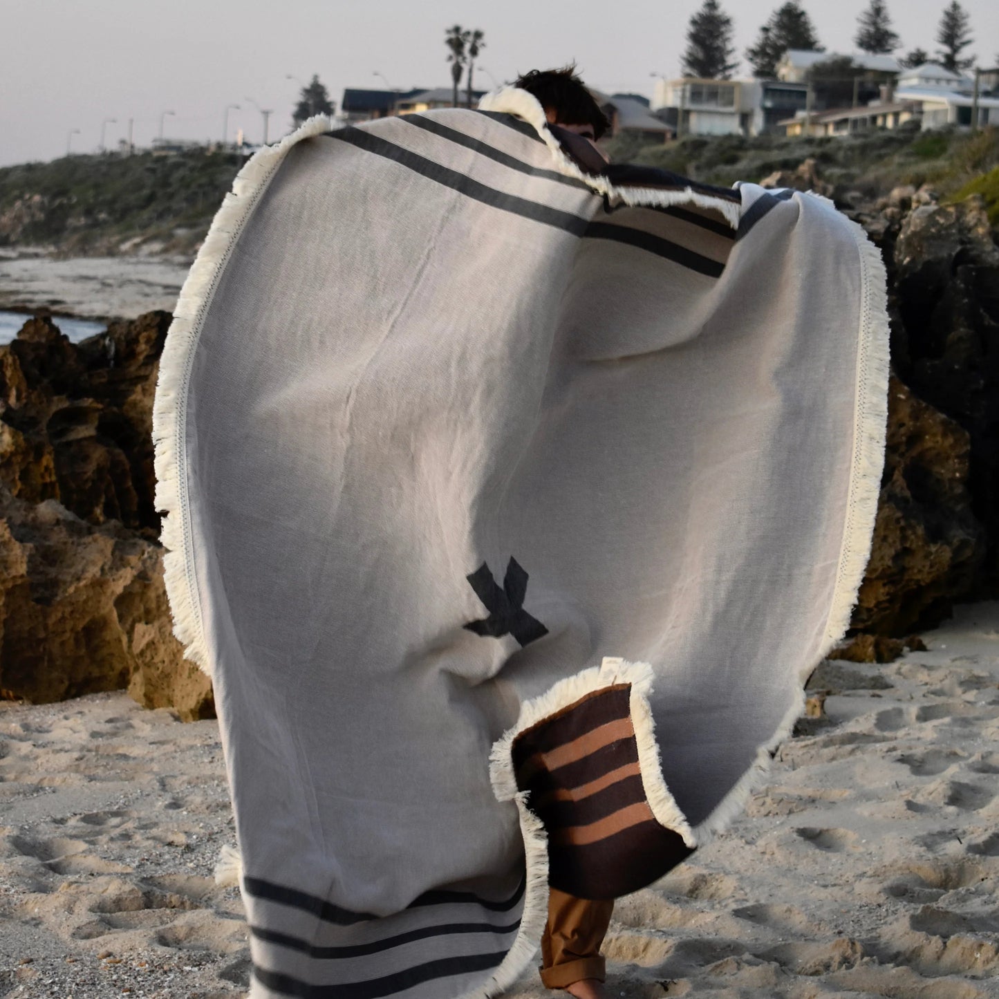 X Marks the Spot picnic blanket in charcoal by Pelli held up by someone standing on the beach with the wind blowing