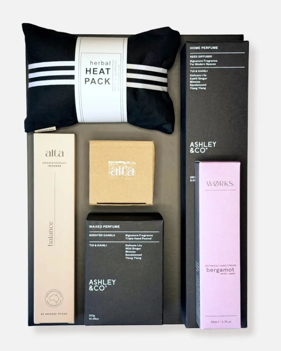 A curated gift set of home fragrance products from Ashley & Co, Alta Melbourne and Works Skincare. The box includes a black and white herbal heat pack, a black home perfume diffuser set in a box, a matching soy candle, a lavender Bergamot essential oil cream, a beige box of natural incense sticks in Balance scent, all in a black re usable gift box, arranged on a neutral background.