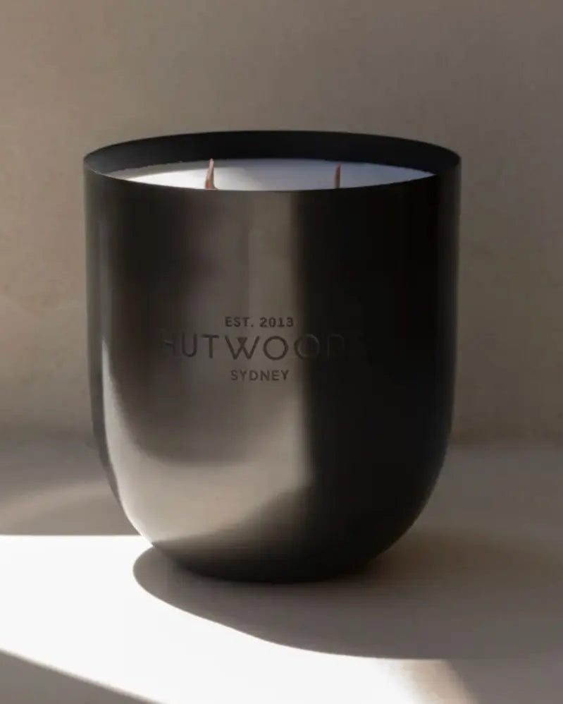 Extra Large Luxe Hutwood candle in dark metal vessel against minimal background in filtered sunlight.. 