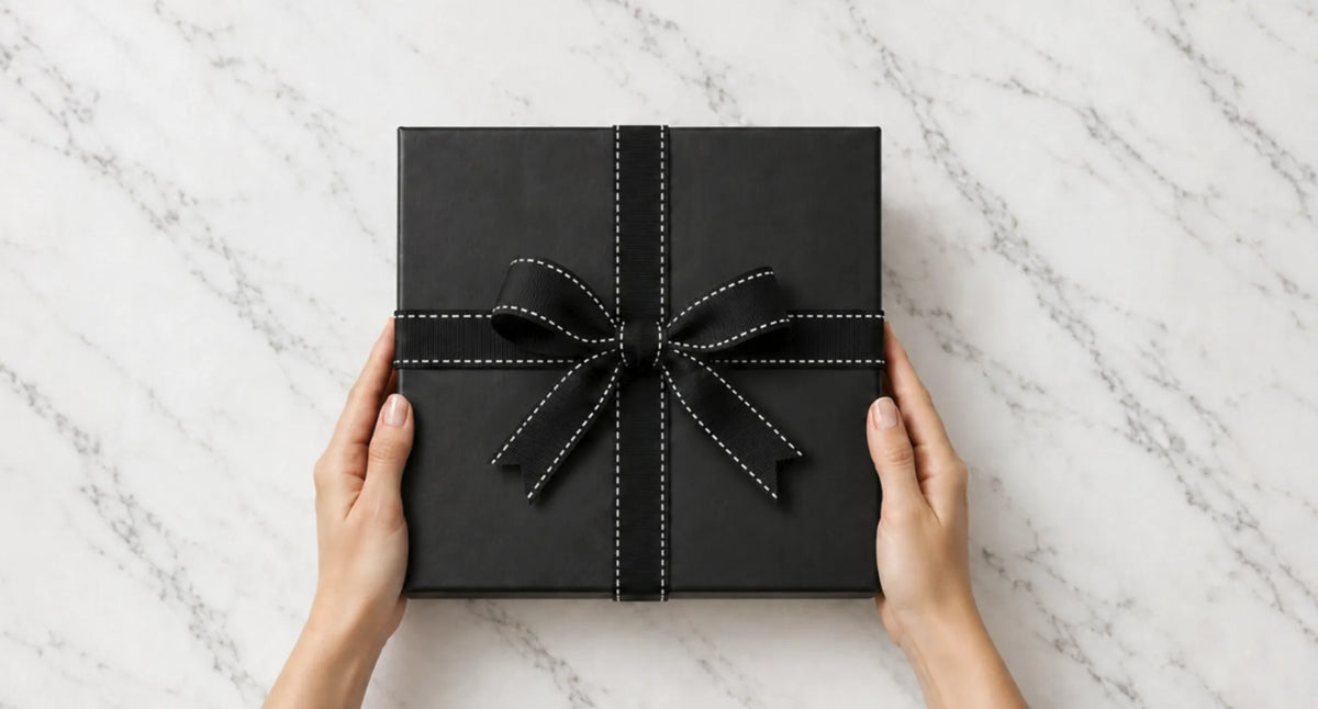 Hands holding a gift wrapped in black paper with a black stitched ribbon bow on a marble bench, wrapped by Hunted + Co. in Sydney
